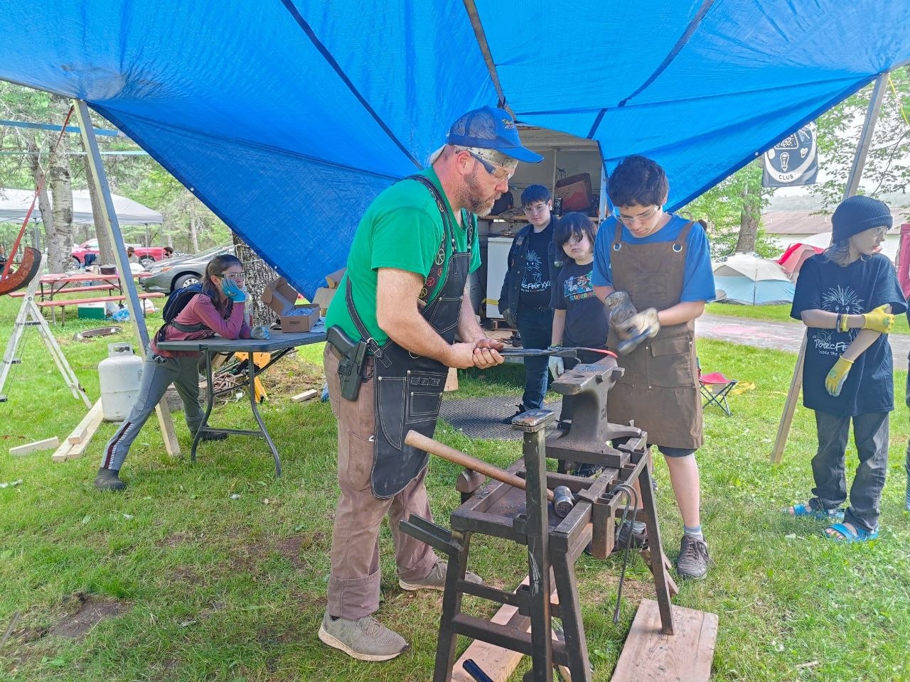 Jay teaching kids blacksmithing under a blue tarp at a Mancamp event